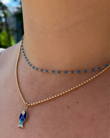 A close-up of a person's neck and collarbone wearing two layered gold necklaces. The top chain is a delicate beaded necklace with small blue beads, and the bottom chain features a gold beaded necklace with a blue and white fish pendant.