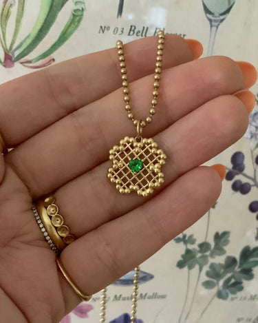A gold beaded chain necklace with a geometric wirework pendant featuring a small green heart-shaped stone, held in a person's hand.