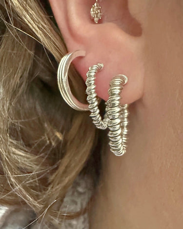 A close-up of a person's ear showcasing a set of textured silver hoop earrings, including one large textured hoop and two smaller twisted hoop earrings, along with a small stud earring.