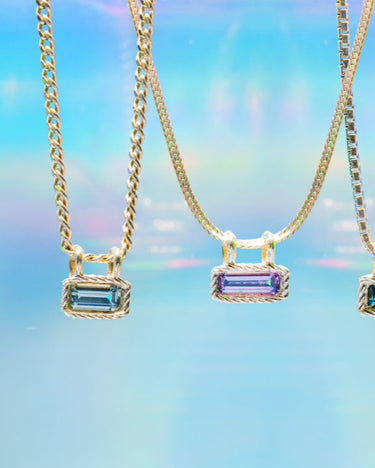 Three gold chain necklaces with rectangular bezel-set gemstone pendants in blue, purple, and teal, hanging against a soft iridescent blue background.