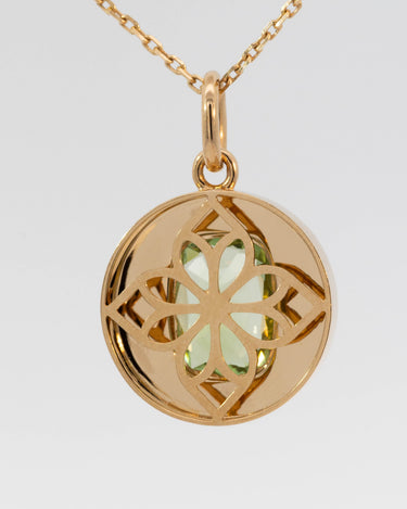 A gold pendant necklace featuring a circular, openwork geometric design with a central light green gemstone, suspended from a gold chain against a light gray background.