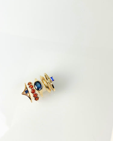 A gold-plated ring with multiple stacked elements, including a star-shaped piece with red and blue stones, a central oval blue stone, and a spiral gold band with a small blue stone, shown on a plain white background.