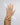A pair of hands with long nude manicured nails, adorned with multiple delicate gold rings featuring small colorful gemstones, set against a soft light gray background with subtle iridescent light leaks.