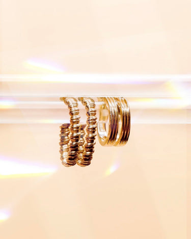 A pair of gold-toned beaded hoop earrings hanging from a clear display bar against a warm cream background with soft light flares.