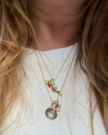 A close-up of a person wearing layered necklaces over a white ribbed top. The necklaces include gold-toned chains with charms, including a small red heart, a gold horse, and a round silver-toned coin pendant.