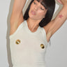 A woman wearing a ribbed white cropped tank top with two prominent gold buttons on the front, paired with light blue sequined jeans, posing with her arms raised against a plain light gray wall.