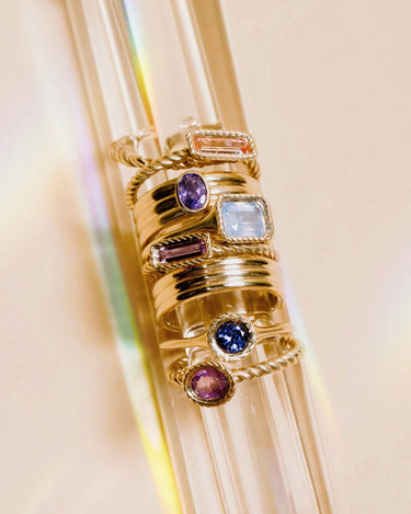 A stack of gold rings with various colored gemstones displayed on a clear glass cylinder against a warm beige background.