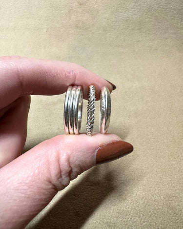 A hand with brown nail polish holds three silver rings against a textured beige background. The rings include a multi-band design, a twisted rope pattern, and a smooth band.